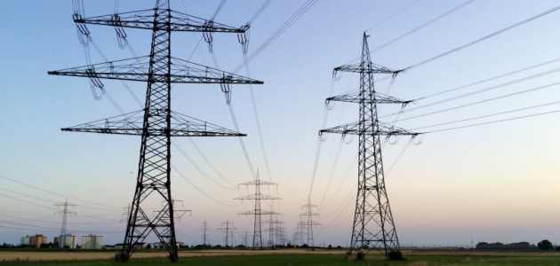 Mehrere große Strommasten mit dazwischen laufenden Kabeln stehen in einer ebenen Landschaft; unten links im Bild sind Wohnhäuser erkennbar.