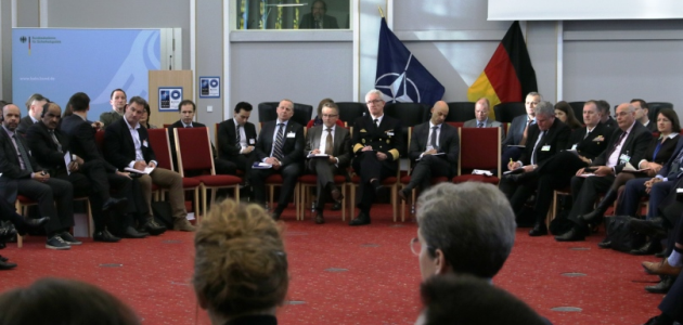 A large group of both uniformed and business-dressed people are sitting in a large circle of chairs within the Federal Academy's Historic Hall.