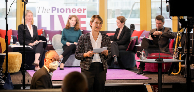 Jana Puglierin spricht mit einem Blatt Papier in der Hand vor einem rundlich angeordneten Diskussionspodium mit vier sitzenden Beteiligten stehen zur betrachtenden Person.