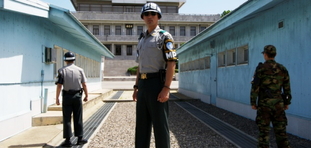 Uniformierte Soldaten mit Helmen und Sonnenbrillen stehen zwischen zwei hellblauen Barackengebäuden und blicken auf ein dahinter aufragendes großes Gebäude.