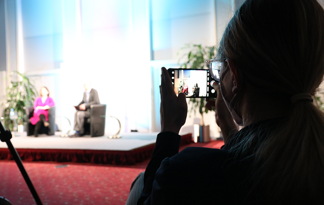 Eine junge Frau fotografiert zwei nur schehmenhaft auf einer Bühne vor einer Pressewand erkennbare Personen