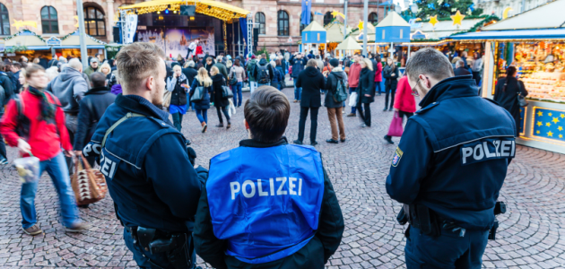 Drei Polizisten stehen bei Tag auf einem Weihnachtsmarkt während zahlreiche Passanten vorbeigehen.