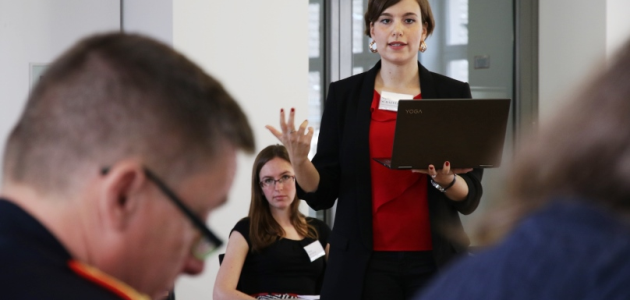 Zwei Frauen blicken zu einem General, der im Vordergrund sitzt; eine der Frauen sitzt; die andere steht, hält ein Notebook in der Hand und spricht gestikulierend.