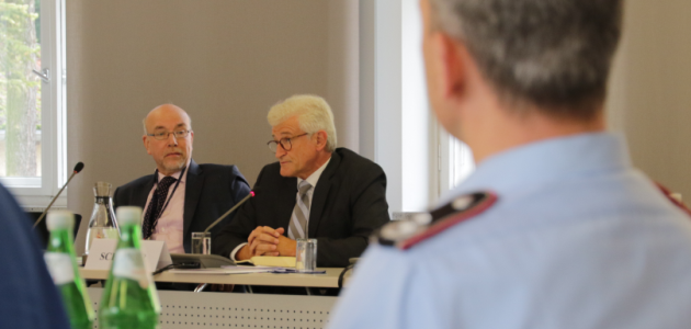 Zwei Herren im Anzug sitzen hinter Mikrofonen an einem Konferenztisch; der links im Bild blickt zu seinem Nachbarn; der rechts im Bild blickt in das Plenum.