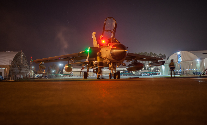 Ein Kampfflugzeug vom Typ Tornado steht bei Nacht auf einem beleuchteten Vorfeld zwischen zwei Hangars.