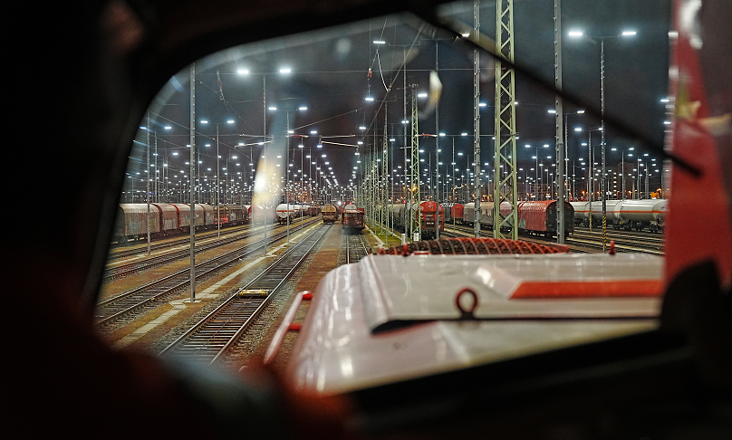 Aus dem Fahrstand einer Lokomotive fällt der Blick auf ein Gleisfeld mit zahlreichen Eisenbahnwaggons bei Nacht, das mit Flutlicht beleuchtet wird.
